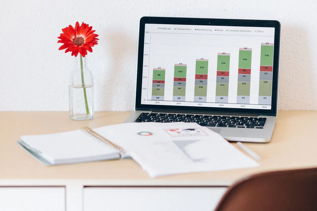 Laptop mostrando gráficos financieros junto a un cuaderno con informes
