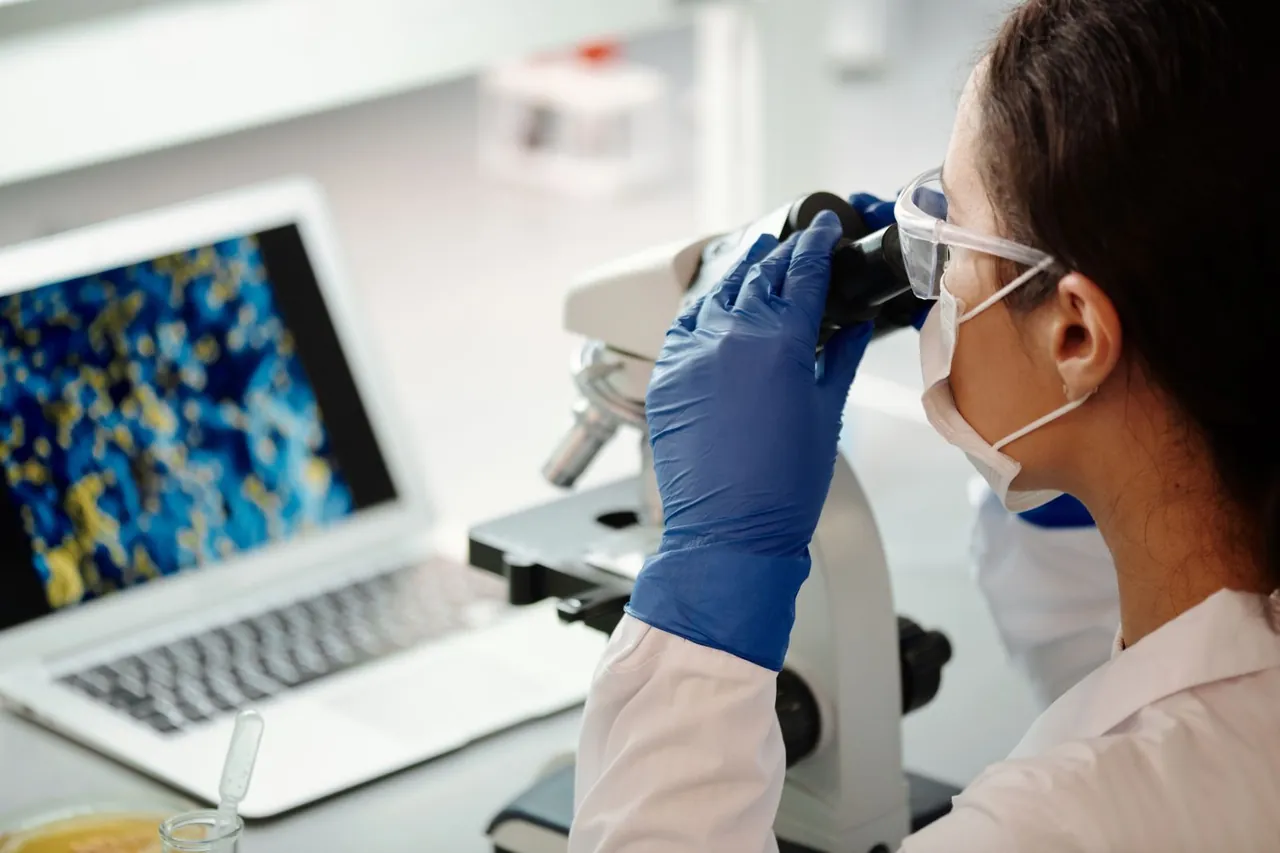 Investigadora con mascarilla y gafas usando un microscopio junto a una laptop